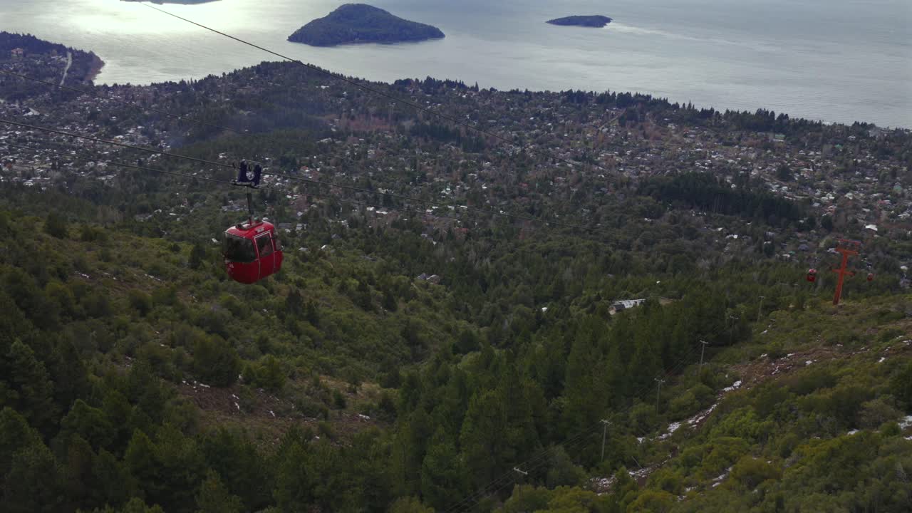 Bariloche ve Nahuel Huapi Gölü'nün panoramik manzarası eşliğinde, ağaçlık bir yamaç üzerinde hareket eden kırmızı kabinli teleferik.