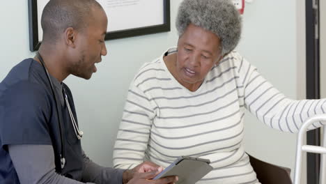 african american male doctor using tablet and talking with senior woman in hospital, slow motion