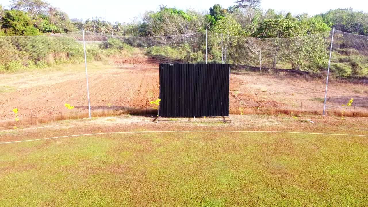 Aerial pull-back shot of a black cricket sight screen on a rural practice field, revealing a boundary line, green grass, and protective netting against a backdrop of tropical trees