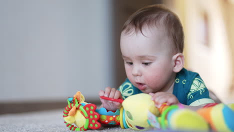 bright toy in the hands of a baby lying on the carpet. focused toddler boy playing with a toy indoors. blurred backdrop.