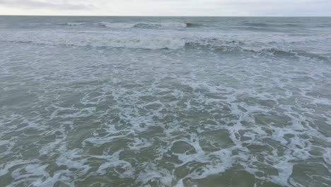 aerial establishing view of big stormy waves breaking against the white sand beach, overcast day, seashore dunes damaged by waves, coastal erosion, climate changes, wide low drone shot moving forward