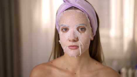 young woman with a face sheet mask and headband looking at camera