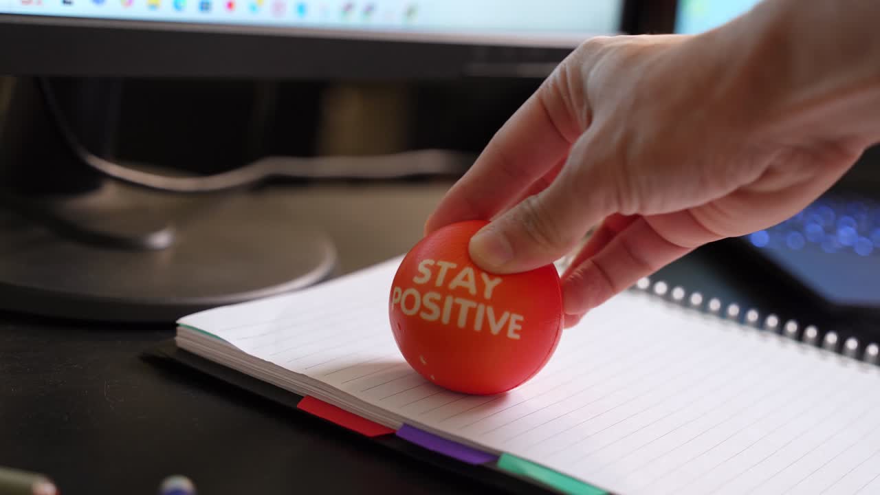 Hand places “Stay Positive” stress ball on notebook at desk. Great for workplace wellness, mental health, daily motivation, and lifestyle content.