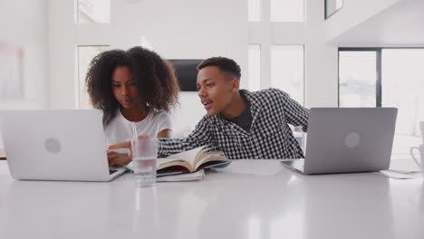 Black-teenage-boy-helping-his-sister-while-they-sit-at-home-using-laptop-computers,-panning-shot