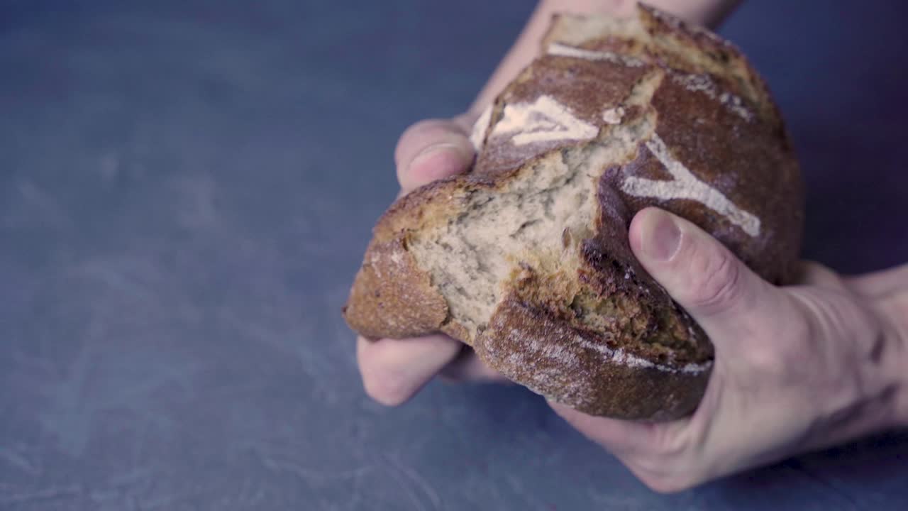 Men's hands break a loaf of round warm bread in half.