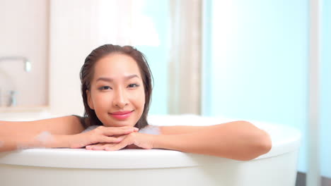 close-up of a smiling young woman leaning her chin and arms along the edge of a large stand-alone bathtub