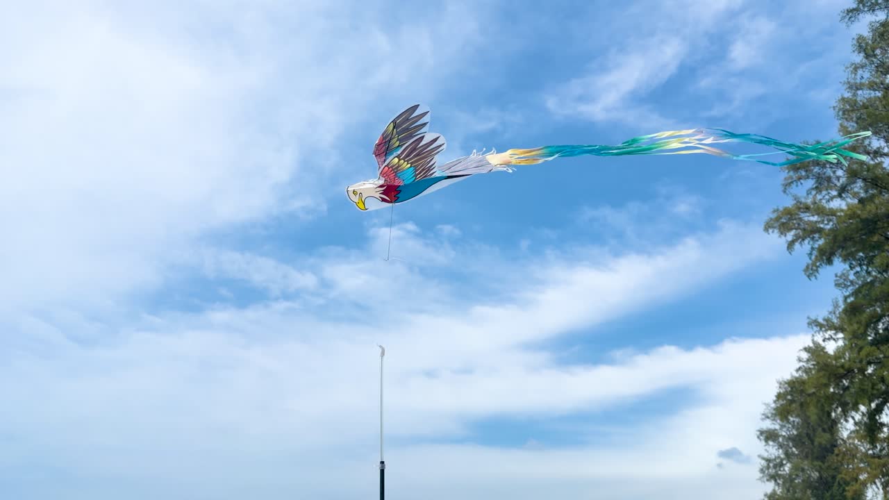 A vibrant bird shaped kite with long streamers flies against a bright cloudy sky