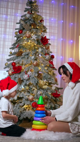 happy mother and little son in santa caps sit at the christmas tree facing each other and talking. cozy festive evening. mom and kid playing with pyramid. vertical video.
