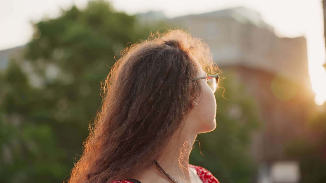Young woman with curly hair glasses backlit by sunset, closeup portrait in urban park, contemplative gaze and soft bokeh, warm rim light on hair, casual red dress with necklace, reflective mood