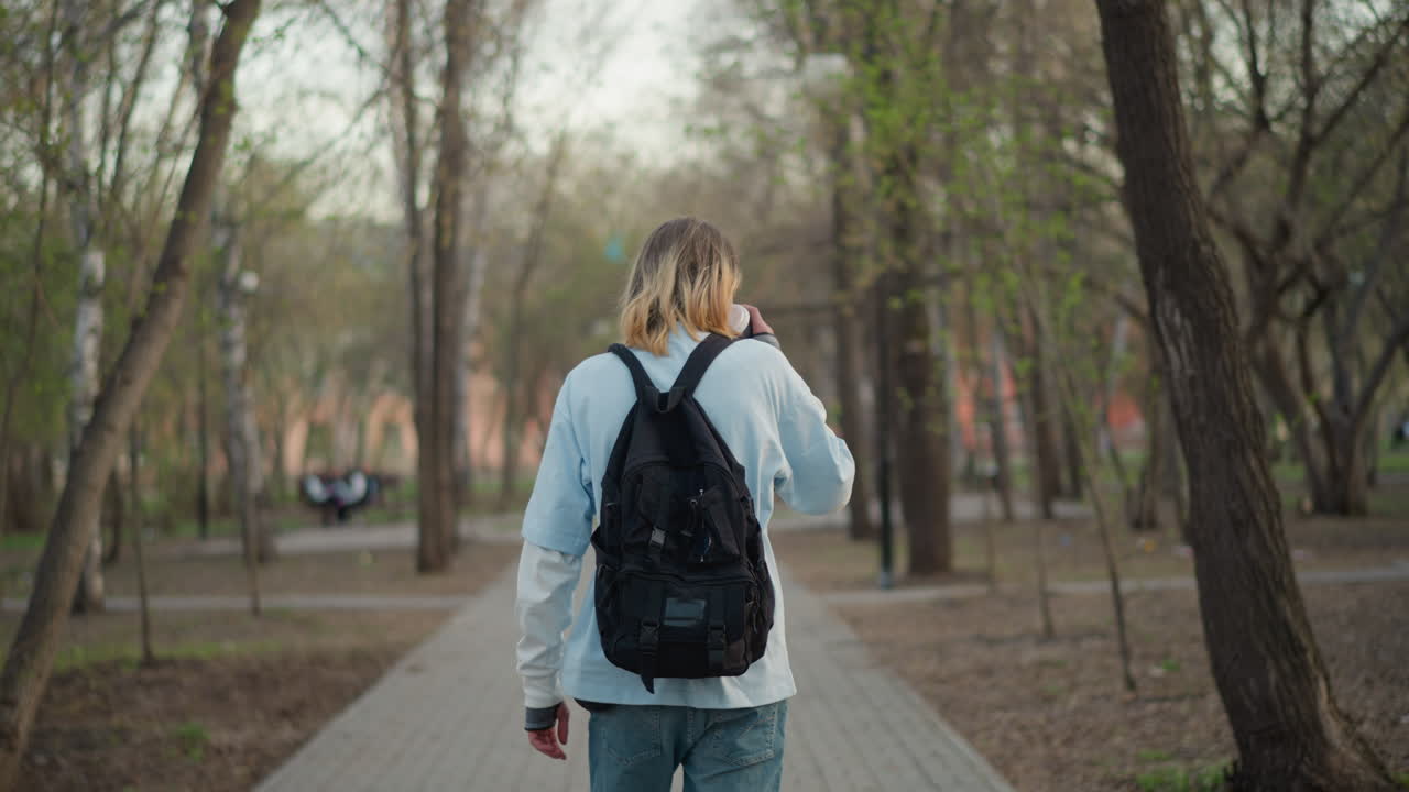 rear view white walker with backpack on paved park path, steady stride past trees and quiet benches, reflective commuter heading through woodland fringe toward city, muted spring tones