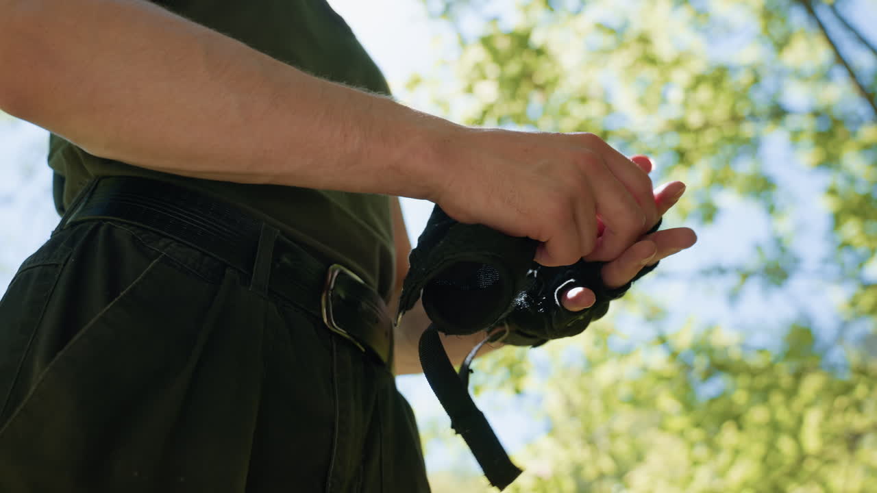 Waist view of tourist pulling off glove to reveal red knuckles, wearing dark cargo pants and belt, hands clad in fingerless tactical gloves, set against backdrop of sunlight and summer sky