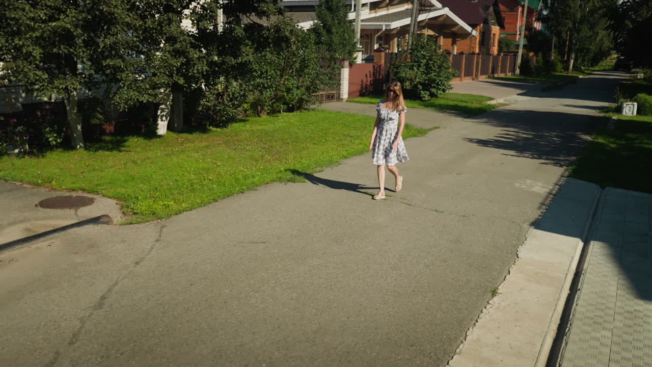 Woman in summer dress walks casually along quiet residential street, swinging arms and clapping hands together in relaxed mood, trees, lawns, and buildings line sidewalk under warm daylight