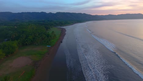 aerial drone shot slowly lowering down towards a sandy beach at sunrise in costa rica over calm rolling waves hitting the shore