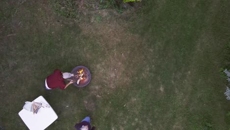 Aerial-birdseye-view-of-a-person-putting-wood-into-a-campfire-with-a-table-nearby-and-some-adults-and-kids-walking-or-running-nearby