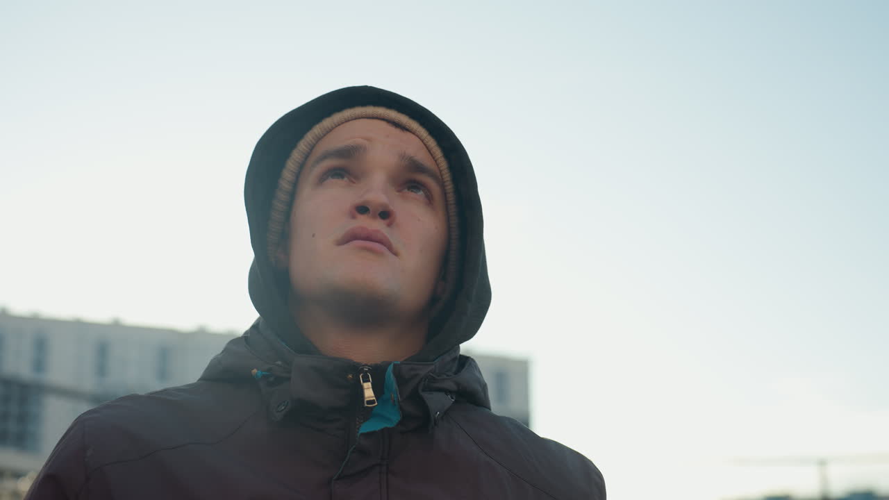 hombre con chaqueta y gorra corriendo en una arena deportiva al aire libre con expresión enfocada, contra un fondo urbano con malla de alambre y edificio de oficinas borroso