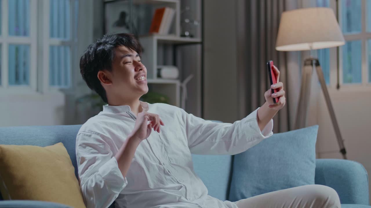 Smiling Asian Man Having Video Call On Smartphone While Sitting On Sofa In The Living Room