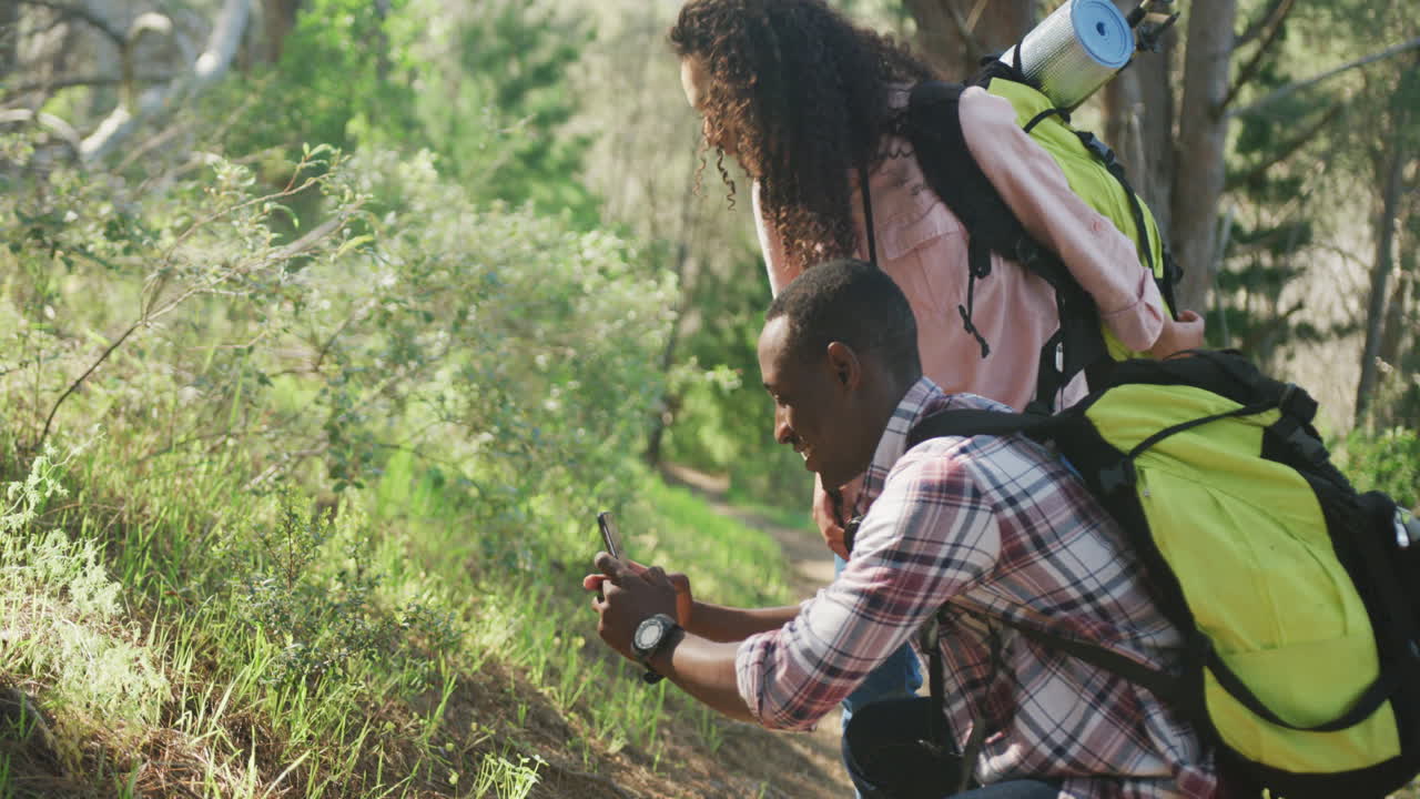 una pareja diversa sonriente tomando fotos con un teléfono inteligente y caminando por el campo