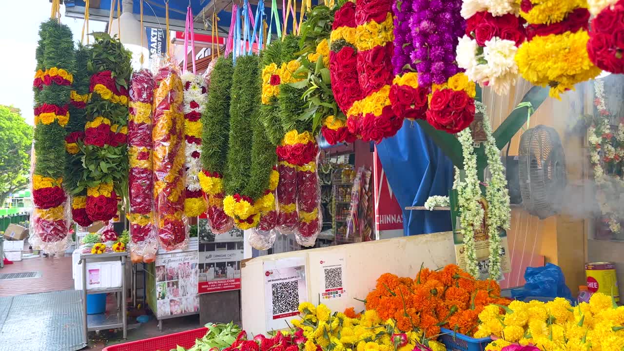 Vibrant flower garlands and loose blossoms displayed at an outdoor market stall in Little India, Singapore. Bright daylight, slow panning camera movement