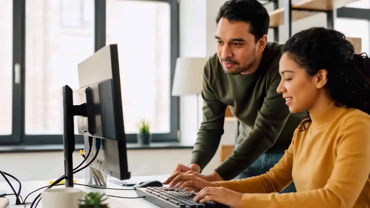 Pointing man in green-sweater guiding woman in mustard typing at desk by windows, showing monitor