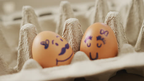 Video-of-close-up-of-two-brown-eggs-with-drawn-faces-in-egg-carton-background