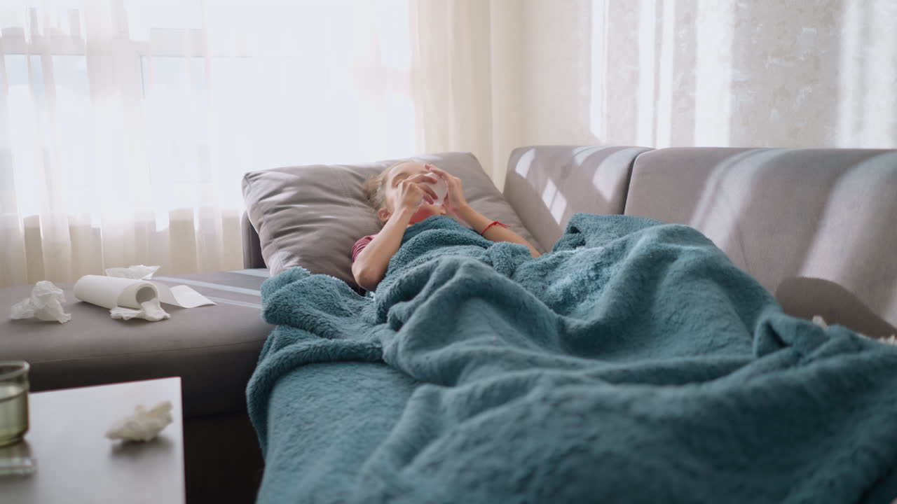 Sick child covered in blanket, sneezing while holding tissue in hand, medication visible on the table, flu symptoms visible at home, tired child resting on couch in a cozy environment