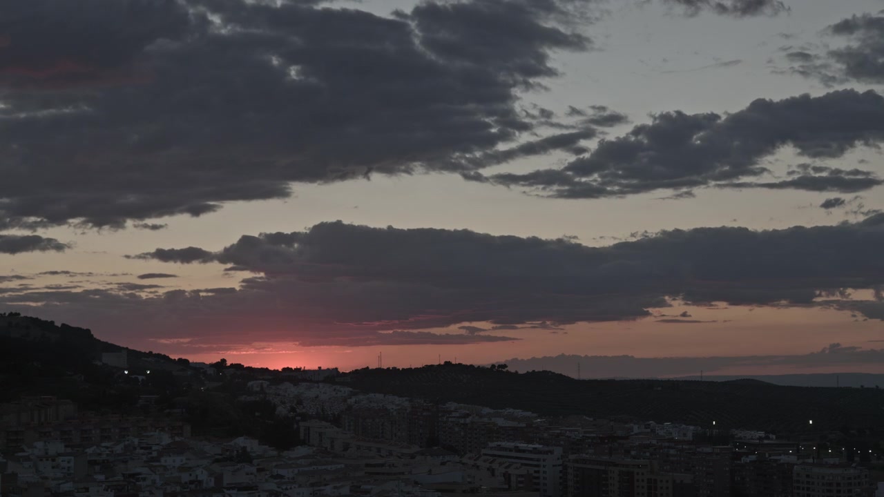 Timelapse de un atardecer sobre el horizonte de una ciudad, mostrando el sol poniéndose detrás de las colinas y el oscurecimiento gradual del cielo nublado.
