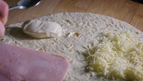 Un-Primer-Plano-De-Una-Cucharada-De-Mayonesa-Rica-Y-Cremosa-Que-Se-Agrega-A-Una-Envoltura-De-Tortilla-De-Jamón-Y-Queso,-El-Condimento-Se-Esparce-Uniformemente-Con-El-Dorso-De-Una-Cuchara