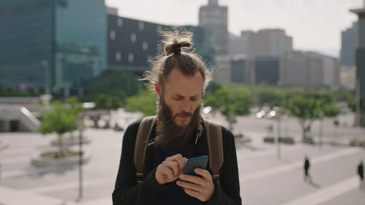 ritratto di un uomo hippie caucasico maturo con la barba che scrive messaggi navigando sui social media usando un'app digitale per smartphone in una città urbana soleggiata