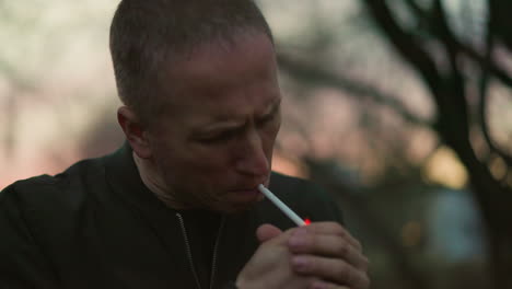 a man in a green jacket lights a cigarette outdoors at dusk, his face illuminated by the red glow of the flame, with blur background of trees