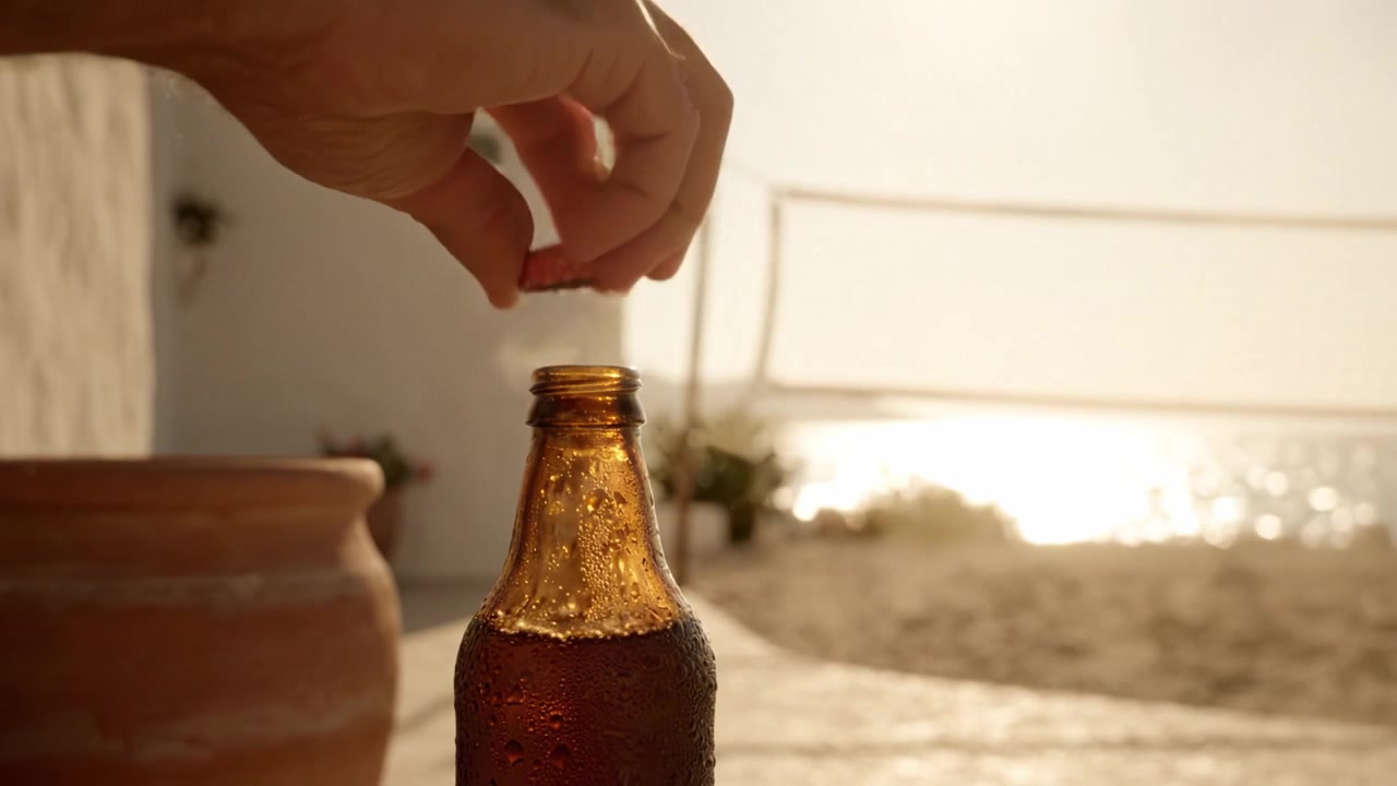 Refresco Gelado: Um Close-up de uma Garrafa de Bebida Gelada Contra um Fundo de Praia Ensolarada com uma Rede de Vôlei ao Longe