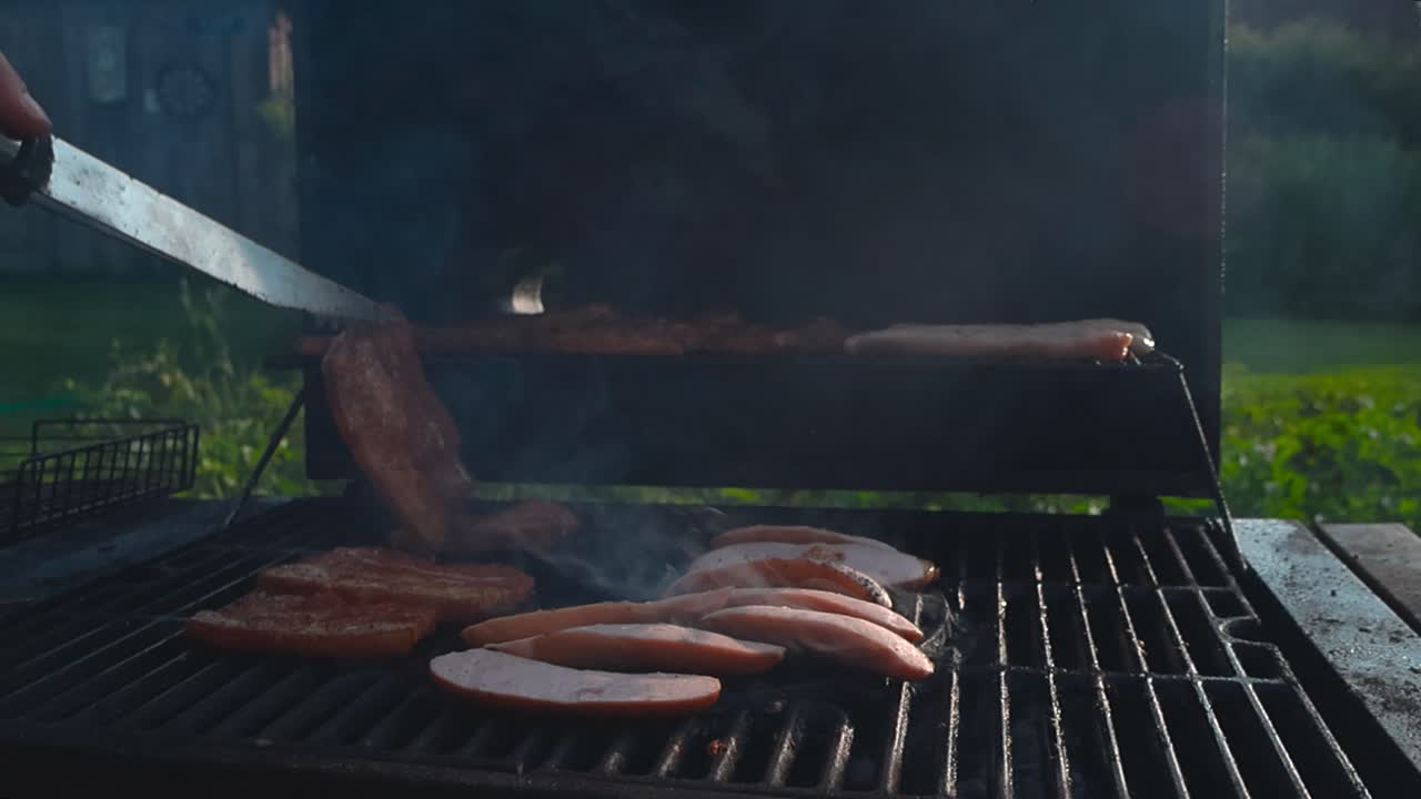 Brown and charred meat and sausages of chicken and beef being grilled on a smoking hot barbecue grill or barbeque during a summer sunny day with white smoke and embers moving in slow motion