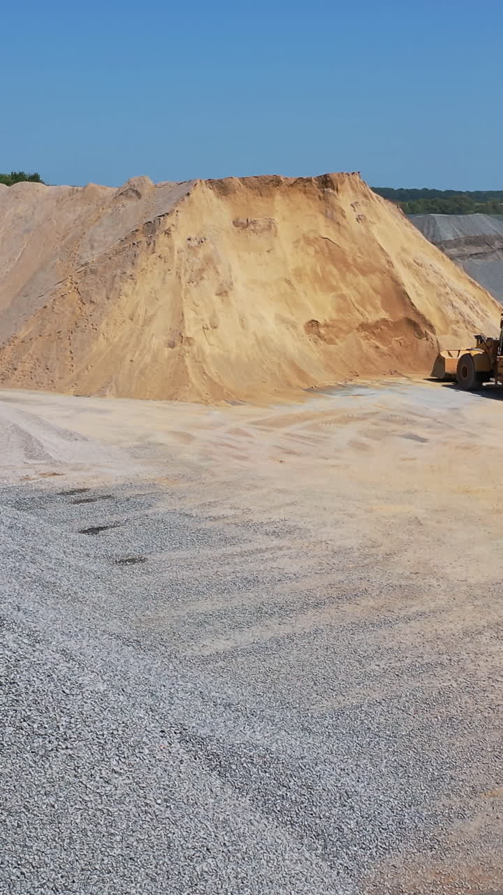 Huge piles of crushed stones and sand outdoors. Bulldozer working on the territory of asphalt plant in a sunny day. Aerial view. Vertical video