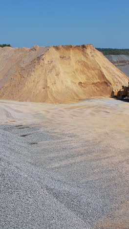 huge piles of crushed stones and sand outdoors. bulldozer working on the territory of asphalt plant in a sunny day. aerial view. vertical video