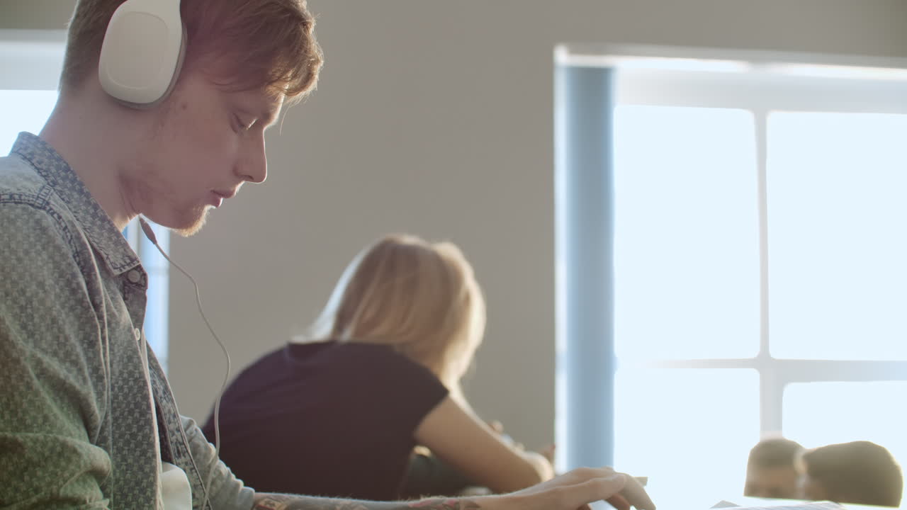 студент, сидящий в университетской аудитории с большой группой людей, сидит в наушниках и слушает музыку и смотрит на экран смартфона. он находится в состоянии спокойствия. перерыв в классе