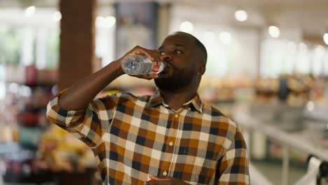 portrait of a man with black skin color in a plaid shirt drinks water from a transparent plastic bottle in a supermarket. shopping break