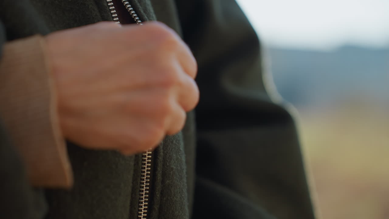 Close up of female hands with red nail polish fastening metal zipper on dark green coat over beige sweater outdoors in daylight showing detail of fashion clothing texture style