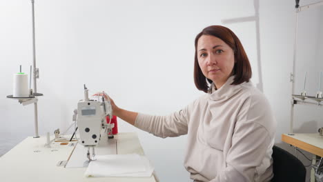 seamstress in light top sitting in bright sewing workshop holding sewing machine with one hand, smiling warmly at camera with relaxed expression surrounded by tailoring tools, and organized workspace