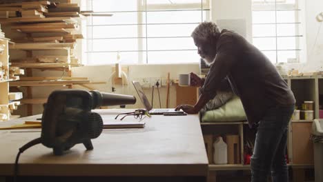 african american male carpenter using laptop while drinking coffee in a carpentry shop