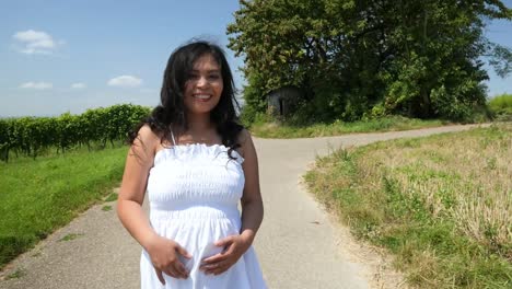 una joven embarazada con un vestido blanco de verano caminando por un camino rural sosteniendo su vientre para una sesión de fotos de maternidad