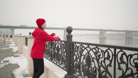 woman in red hoodie and black leggings rests hand on decorative metal fence with gloves, gazing at distant bridge with moving cars, surrounded by snow-covered ground and river