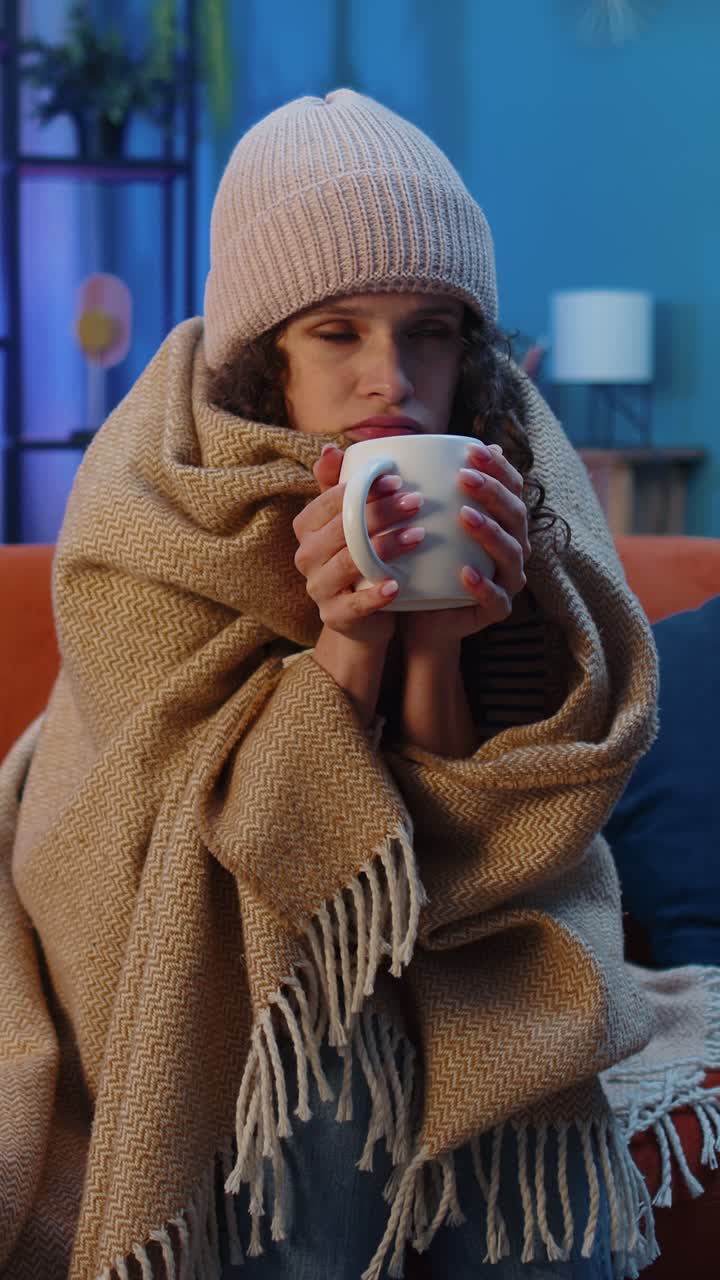 Una donna malata e malata indossa un cappello avvolto in un quadri si siede tremando di freddo sul divano a bere tè caldo