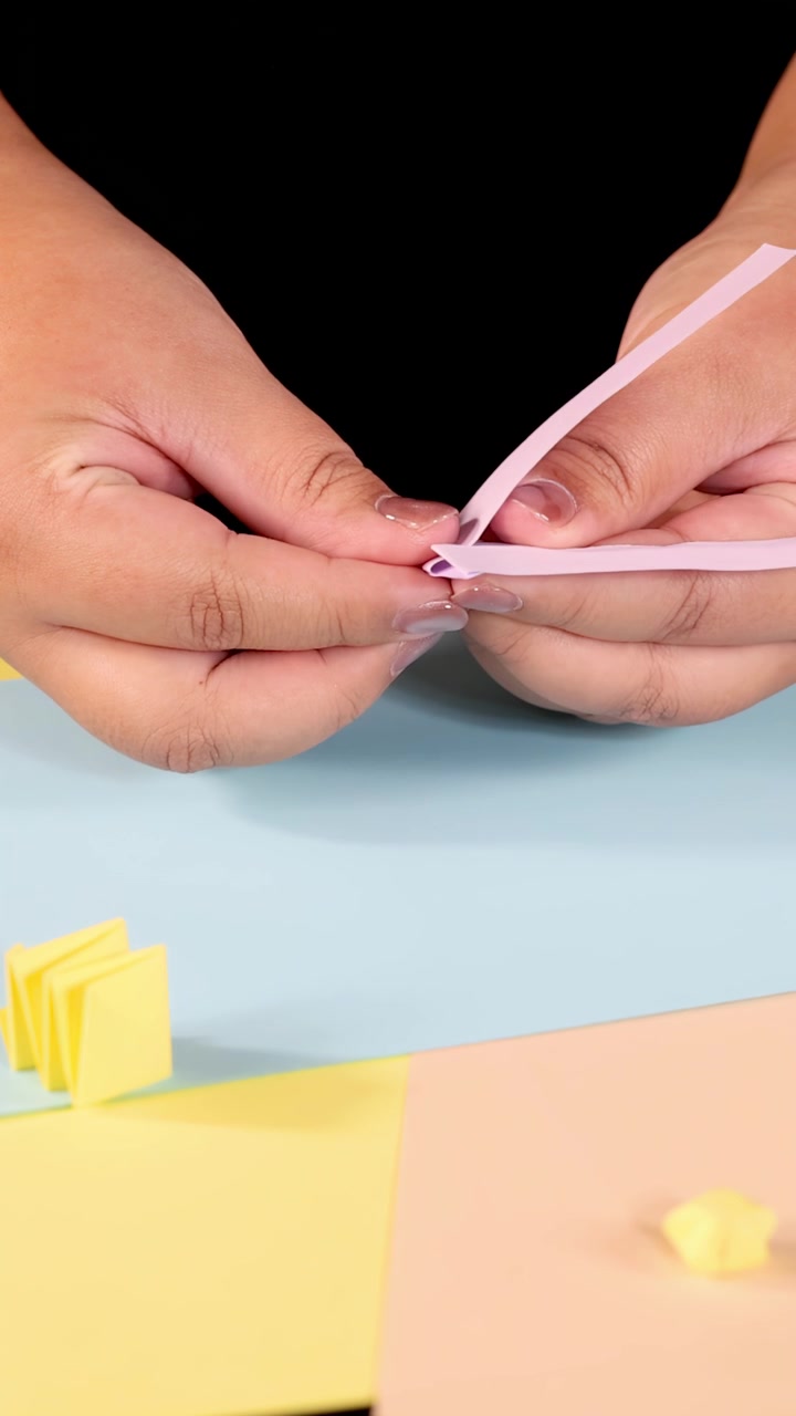Close-up of hands folding pink paper strip on pastel surface, bright lighting, static camera