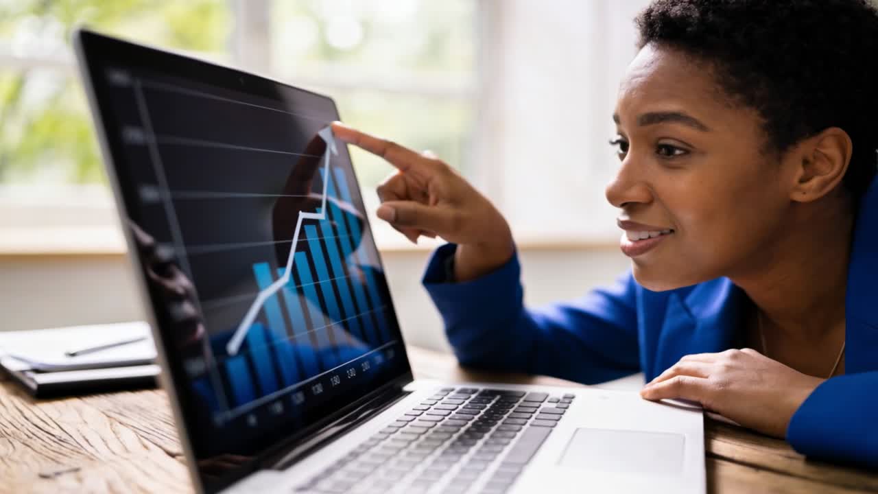 Seeing upward trend on laptop at desk, woman in blazer pointing, smiling, making fist, copy space