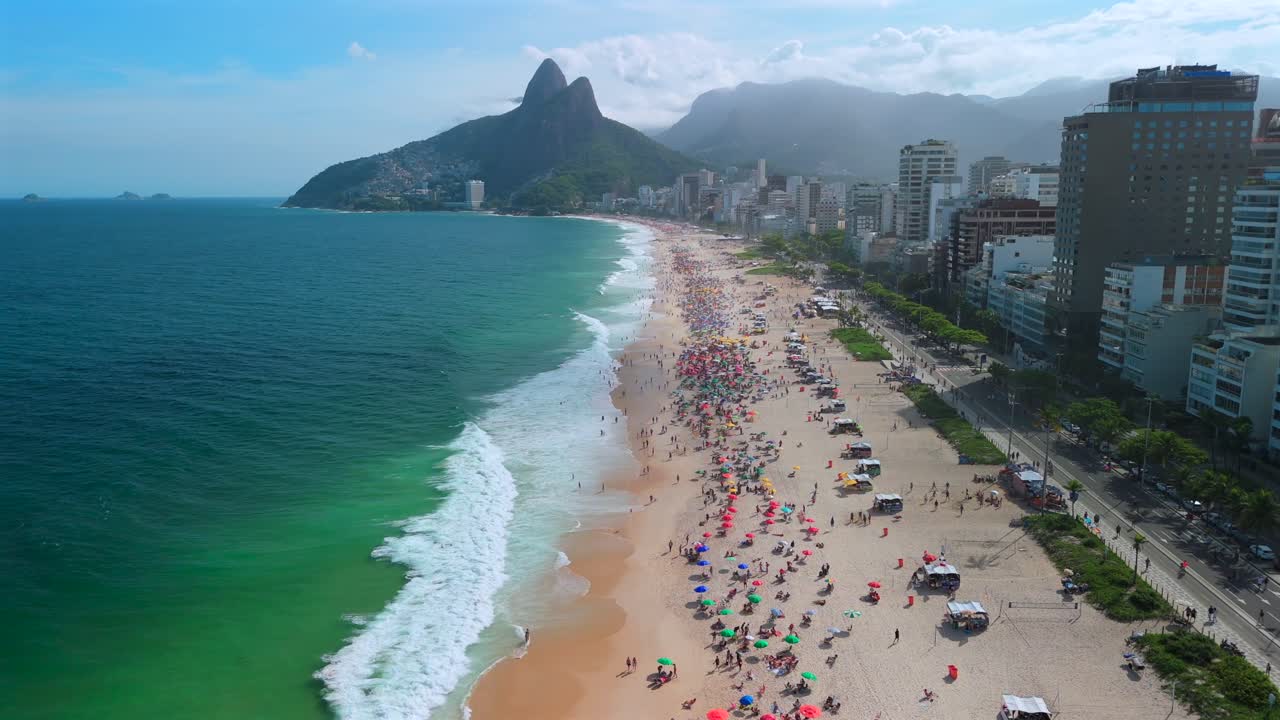 Aerial pull-back reveals umbrella-dotted Ipanema Beach packed with sun-seekers under bright Rio skie