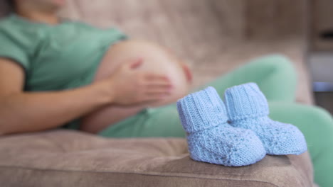 little knitted socks for future baby. blue baby socks on sofa on the blur background of a pregnant woman stroking her belly.