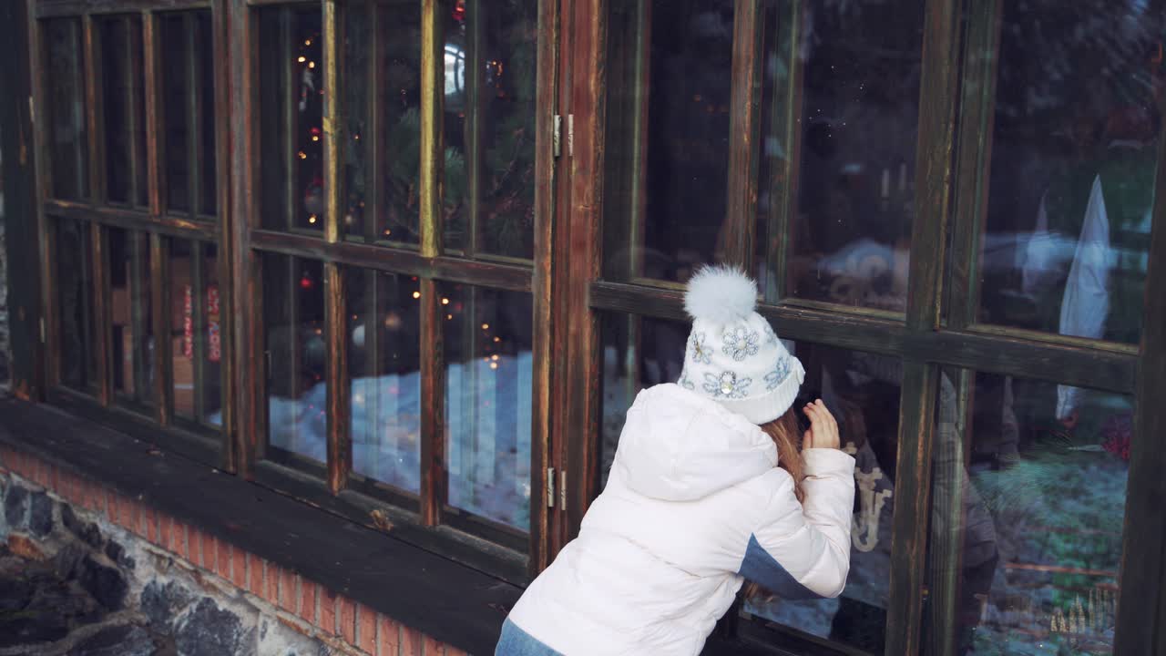 Little girl is looking through the window into the house outdoors. Small child in warm clothes standing near the windows in winter.