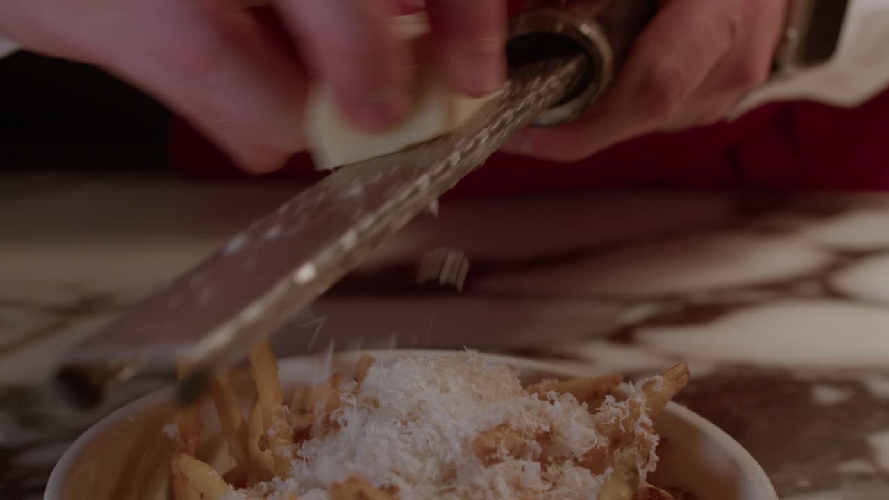 Un primo piano professionale di uno chef che usa una grattugia per aggiungere un generoso strato di formaggio fresco a una ciotola di patatine fritte croccanti su un piano di lavoro in marmo