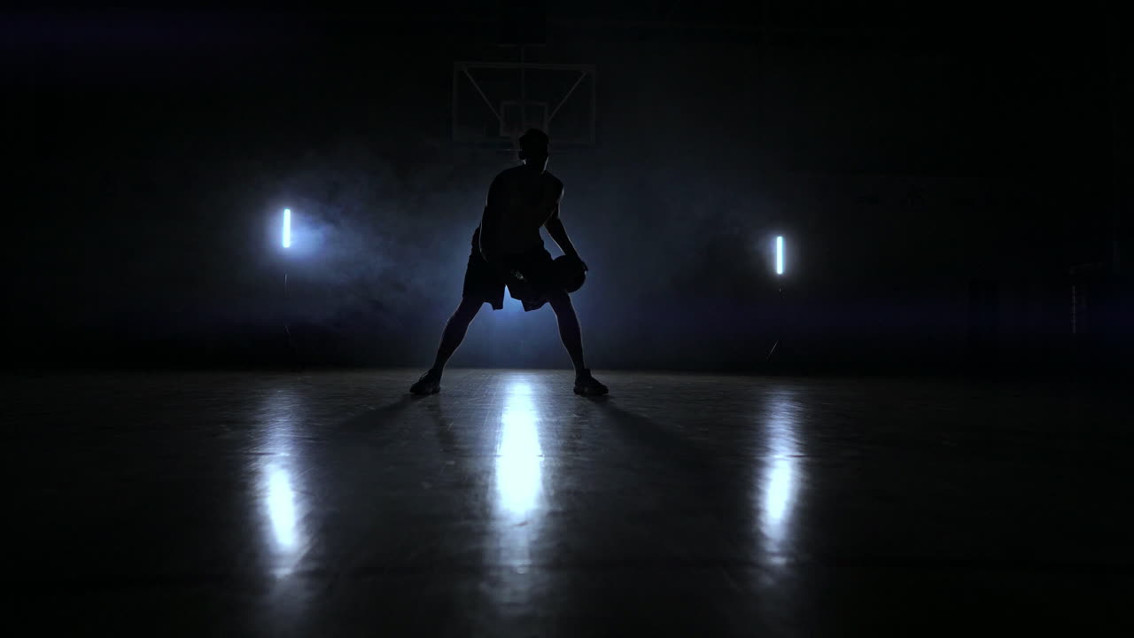 un hombre con una pelota de baloncesto en una cancha de baloncesto oscura contra el telón de fondo de un ring de baloncesto en el humo muestra habilidades de dribbling iluminadas por tres linternas en la luz de fondo