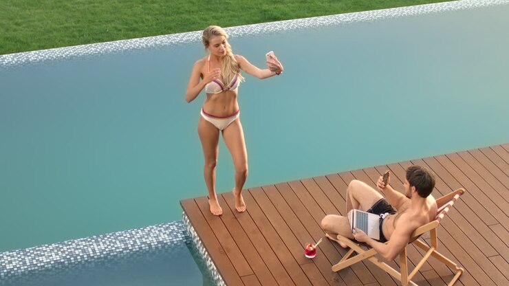 Blonde woman taking mobile selfie near poolside. Couple relaxing near pool
