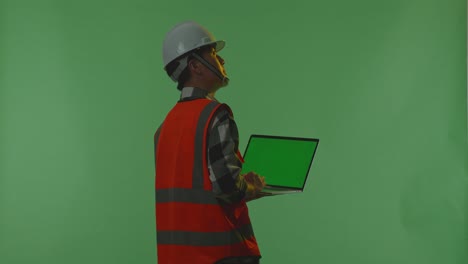 back view of asian male engineer with safety helmet working on a green screen laptop and looking around while standing in the green screen background studio, during sunset or sunrise time